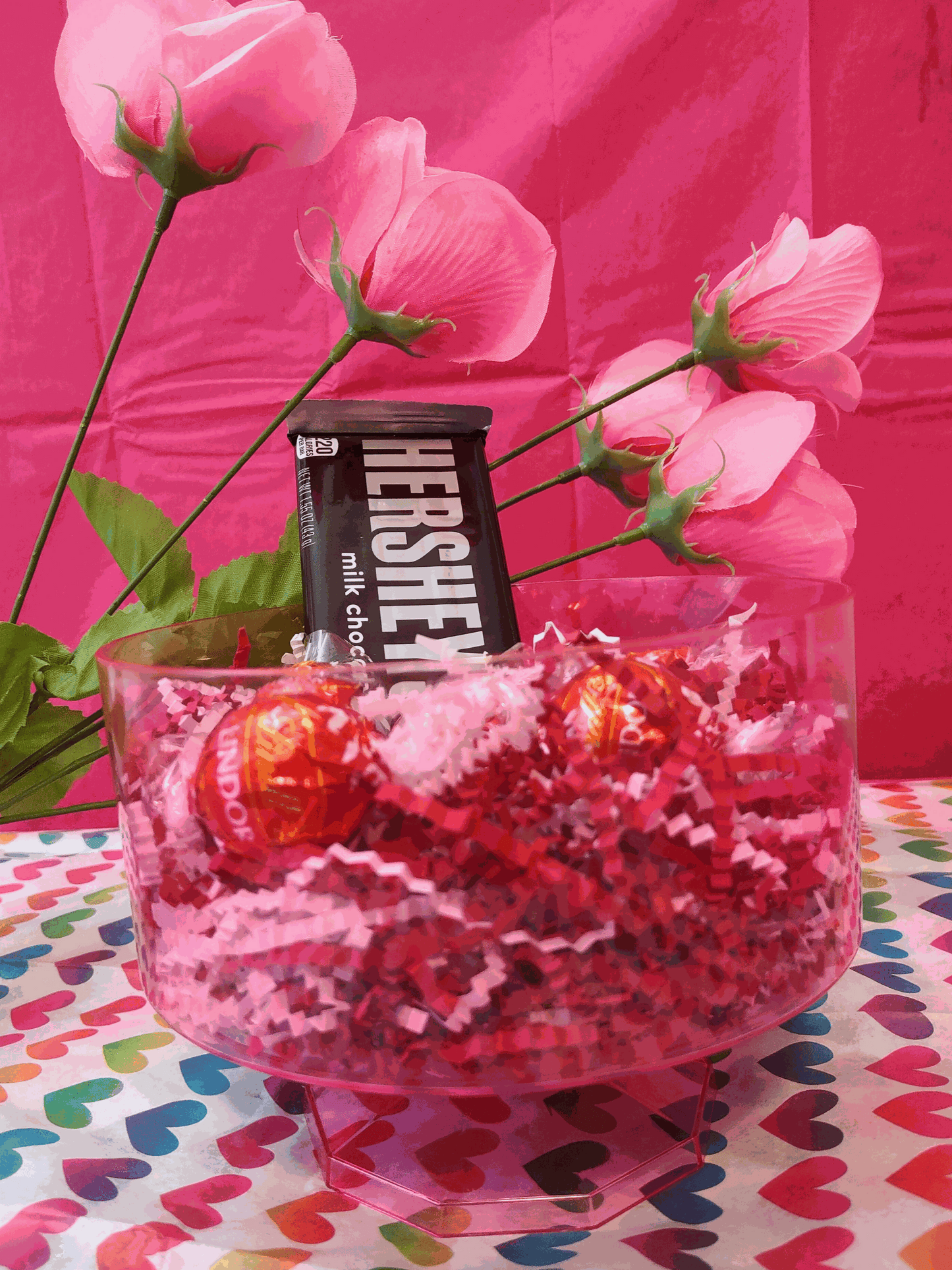 Clear round bowl filled with vibrant red crinkle cut paper, containing a Hershey's milk chocolate bar and two Lindor truffles, against a pink backdrop with pink flowers and a colorful heart-patterned tablecloth.