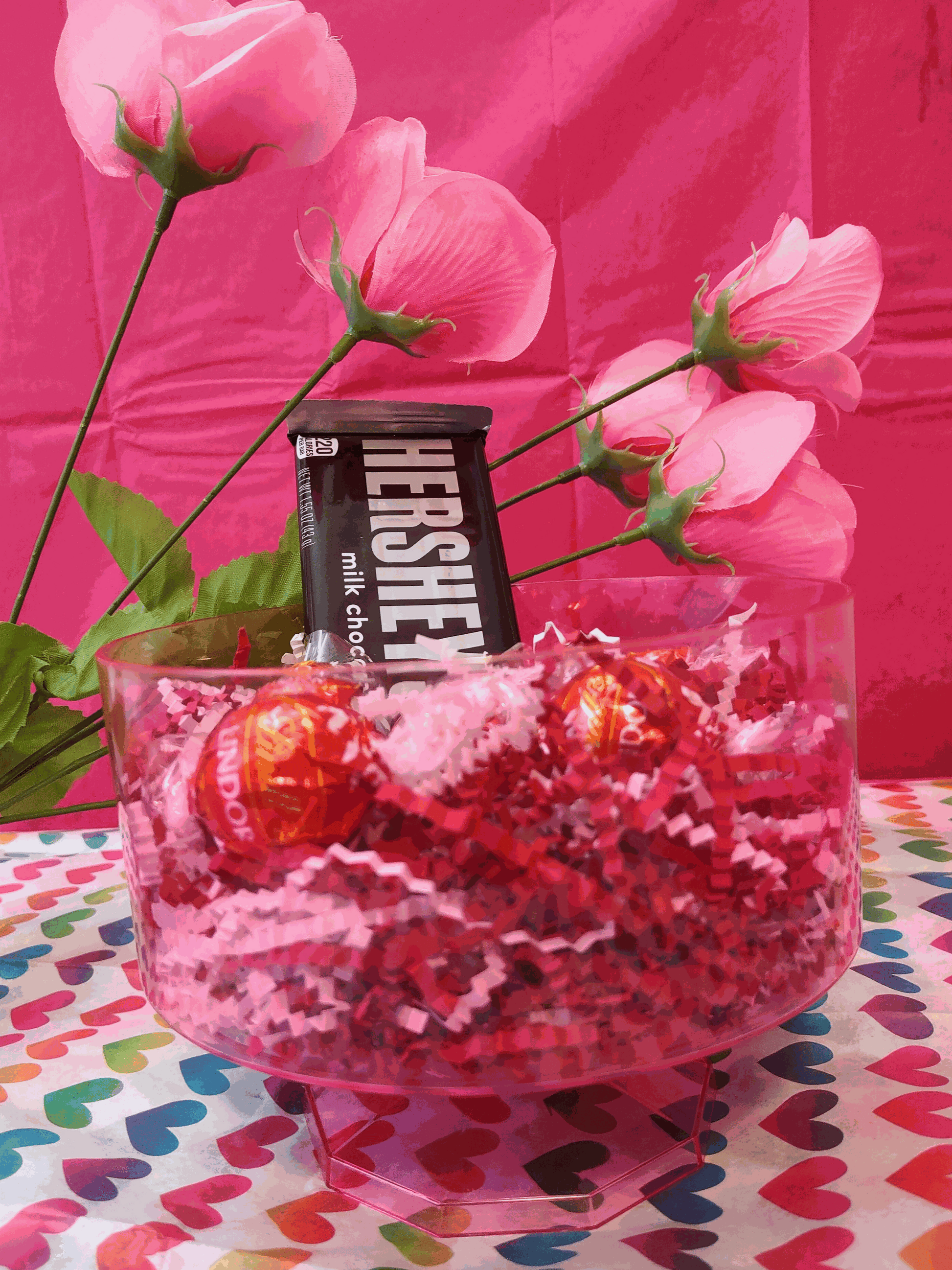 Clear round bowl filled with vibrant red crinkle cut paper, containing a Hershey's milk chocolate bar and two Lindor truffles, against a pink backdrop with pink flowers and a colorful heart-patterned tablecloth.
